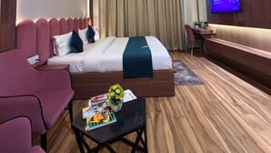 A hotel room with a bed, a desk, a TV, a ceiling fan, and a small table with a plant and magazines. - Infinity Hotel (Kanpur)