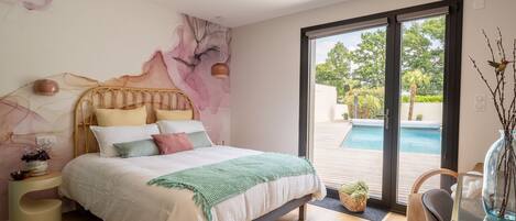 A bedroom with a bed, a nightstand, a chair, a table, and a view of an outdoor swimming pool.