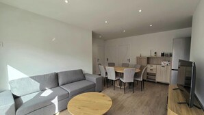 Un salon moderne avec un canapé gris, une table basse ronde en bois, une table à manger avec des chaises, et un coin cuisine équipé d’un réfrigérateur et de placards.