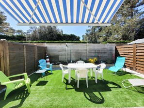 Un patio trasero con una mesa y sillas blancas, un dosel azul y blanco a rayas y una valla de madera.