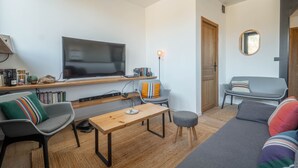 Una sala de estar moderna con una televisión de pantalla plana, una mesa de centro de madera, un sofá gris y un otoman redondo pequeño.