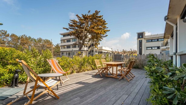Una terraza de madera con mobiliario exterior, un árbol y un edificio al fondo.