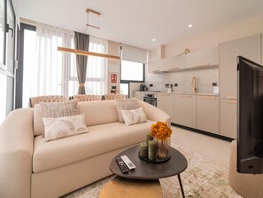 Ein modernes Wohnzimmer mit einem beigefarbenen Sofa, einem Couchtisch mit Blumen und einer Küchenzeile mit hellen Schränken und einem schwarzen Kühlschrank.