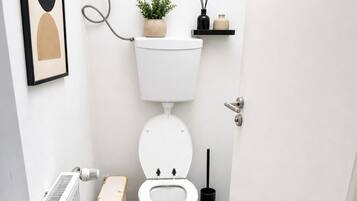 Un baño pequeño y moderno con inodoro blanco, un radiador, un felpudo beige, una planta en maceta y un estante negro con un florero y difusor de cañas.