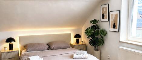 A modern bedroom with a large bed, bedside lamps, a potted plant, and framed artwork on the wall.