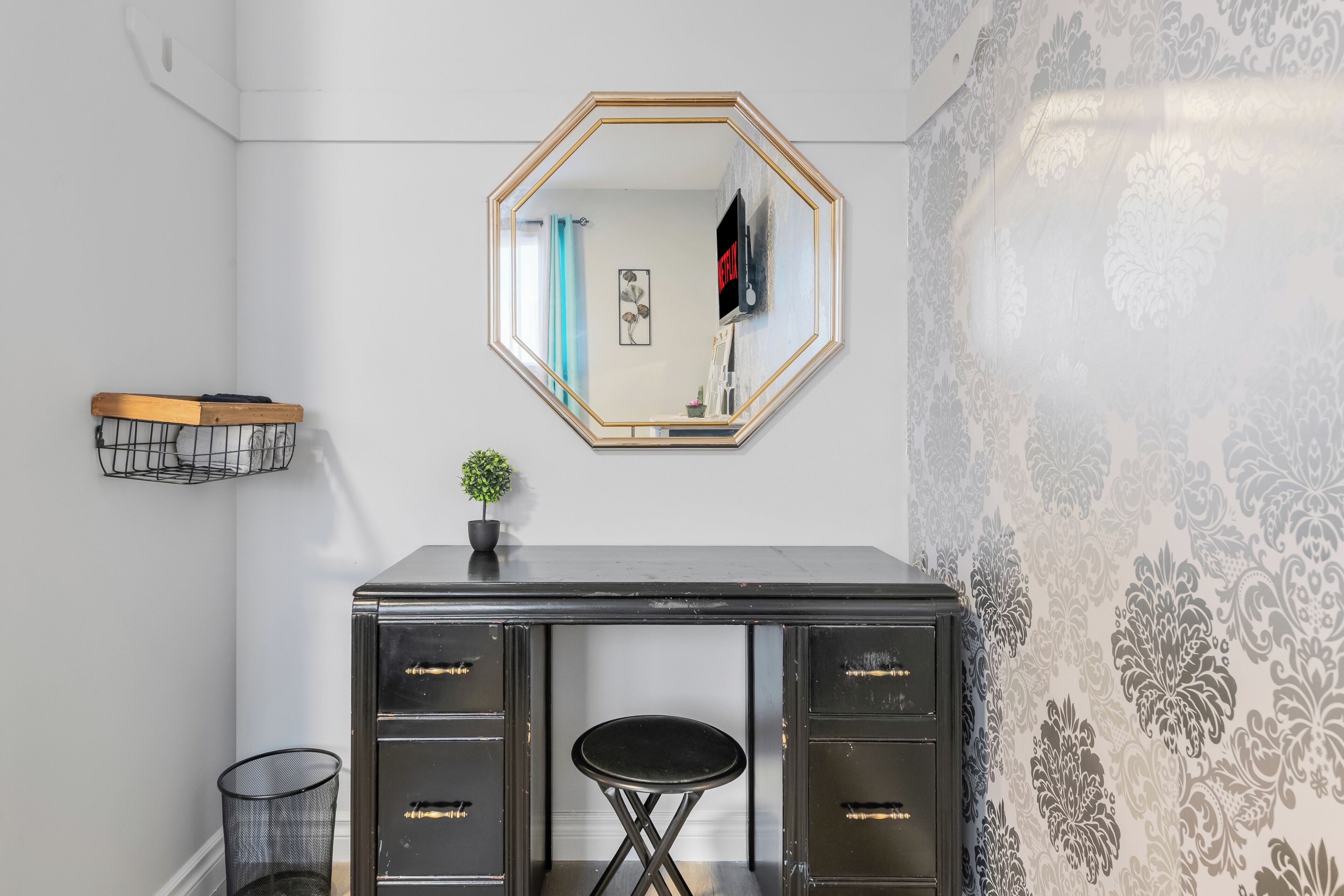 A room with a black desk, a small stool, a mirror, a plant, and a wall with patterned wallpaper.