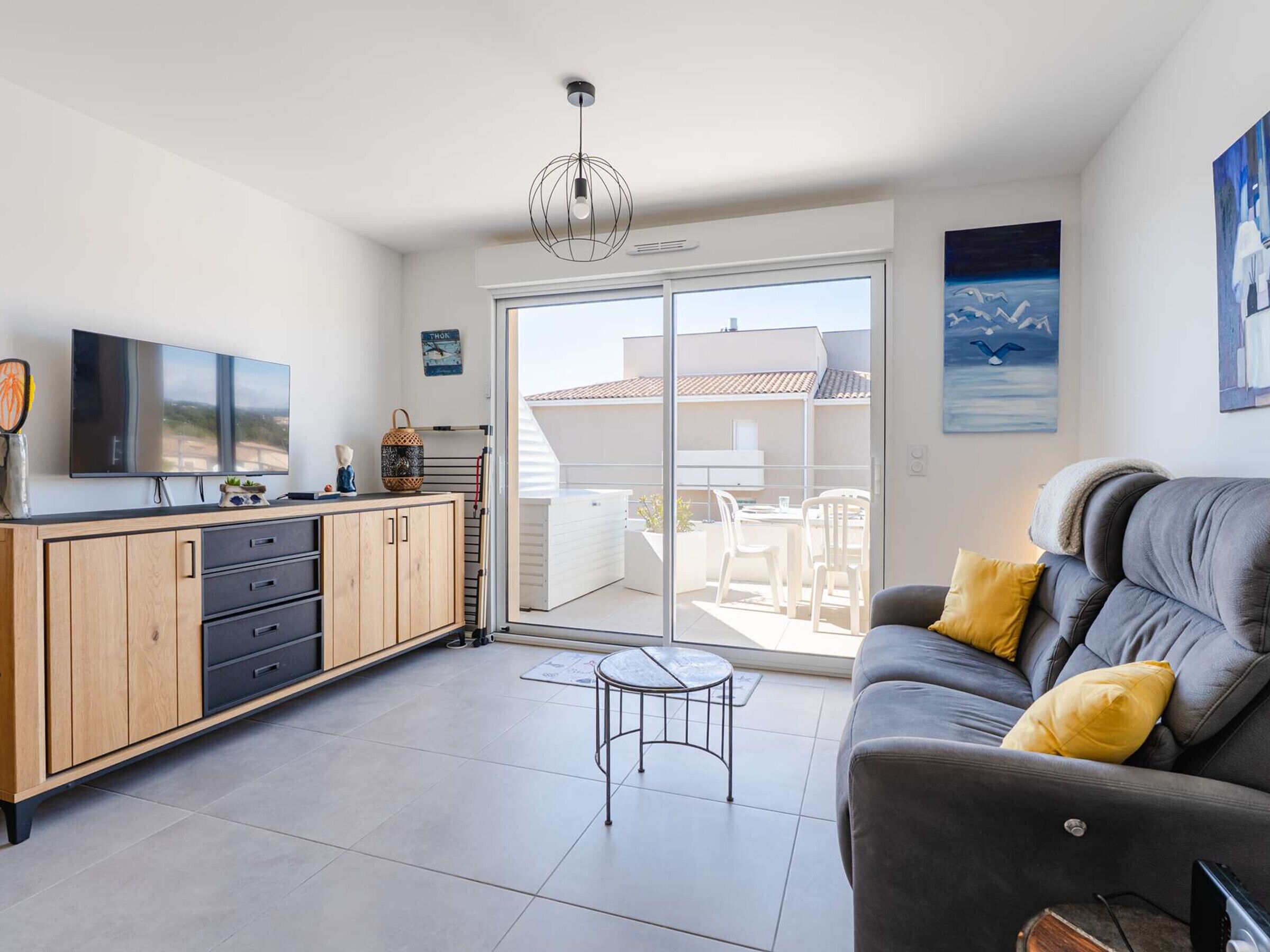 Ein modernes Wohnzimmer mit einem grauen Sofa, einem kleinen runden Tisch, einem Fernseher auf einer Holzkommode und einer Glastür, die auf eine Terrasse im Freien führt.