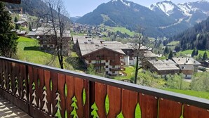Un village de montagne aux bâtiments traditionnels en bois, doté d’un balcon orné d’une balustrade décorative, et offrant une vue sur les collines et les montagnes environnantes.