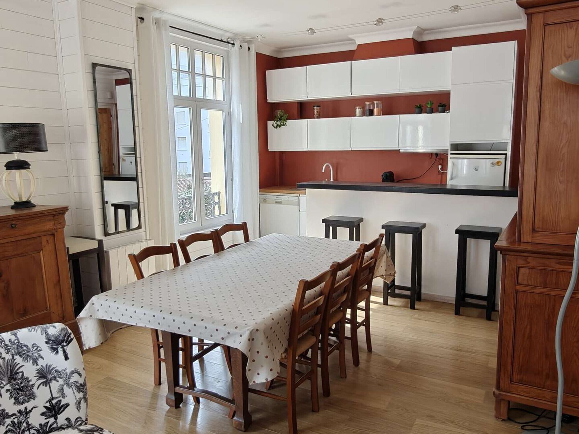 Een eethoek met een tafel en stoelen, een keuken met witte kasten, en een raam met gordijnen.