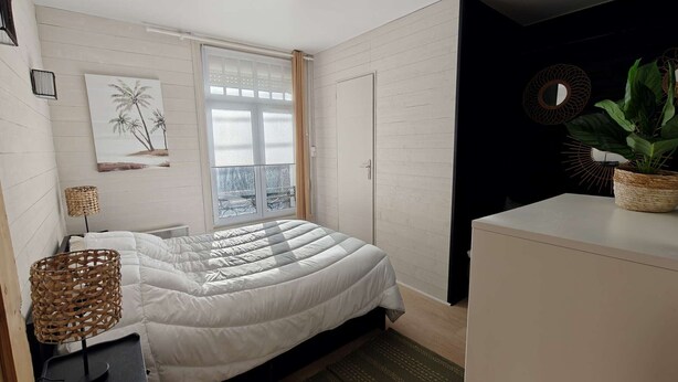 Une chambre à coucher comprenant un lit, une table de chevet, une lampe, une fenêtre avec des stores, une œuvre d’art accrochée au mur, une plante en pot et une commode.