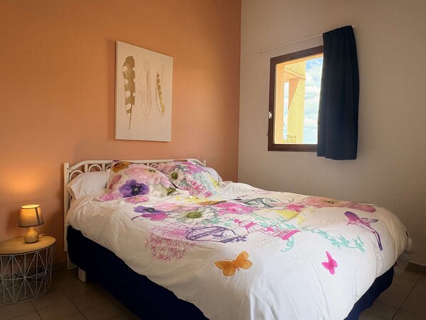 Une chambre à coucher comprenant un lit, une table de chevet, une lampe, une fenêtre avec des rideaux et un tableau accroché au mur.