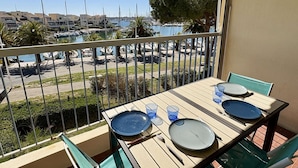 Un balcon avec une table dressée pour deux personnes, donnant sur une marina où l’on peut admirer des bateaux et des bâtiments.