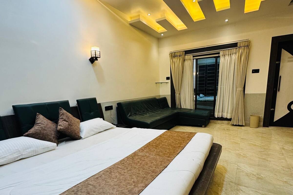 A modern hotel room with a large bed, a sofa, a window with curtains, and a ceiling with recessed lighting.