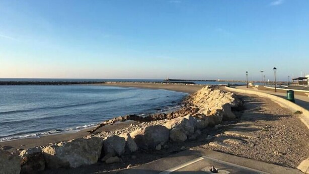 A coastal walkway with a rocky barrier, a clear sky, and a distant shoreline. - House located on a pedestrian street leading to the beach, with private patio. (Saintes-Maries-de-la-Mer)