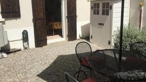 A covered outdoor seating area with a table and chairs, a white door with a window, and a gravel ground. - House located on a pedestrian street leading to the beach, with private patio. (Saintes-Maries-de-la-Mer)
