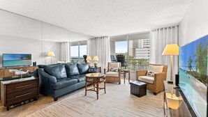A modern hotel room with a large flat-screen TV, a leather sofa, a wicker armchair, a small round table, and a view of the cityscape through large windows. - S Hampton 301 2 Br Oceanview in Kingston, Pool (Myrtle Beach)