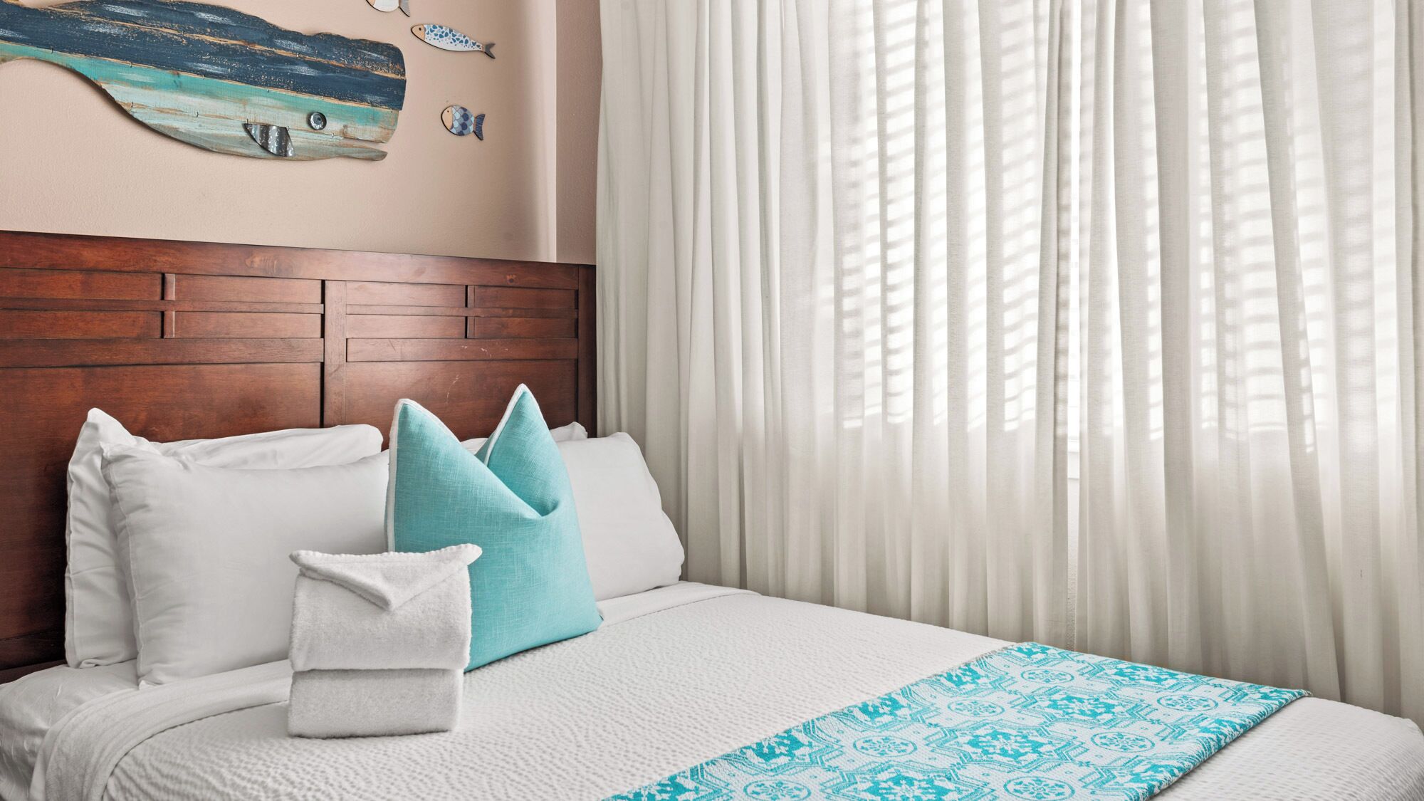 Un lit bien fait, avec une literie blanche et un couvre-lit bleu à motifs, une tête de lit en bois et un mur orné d’une fresque représentant des poissons.