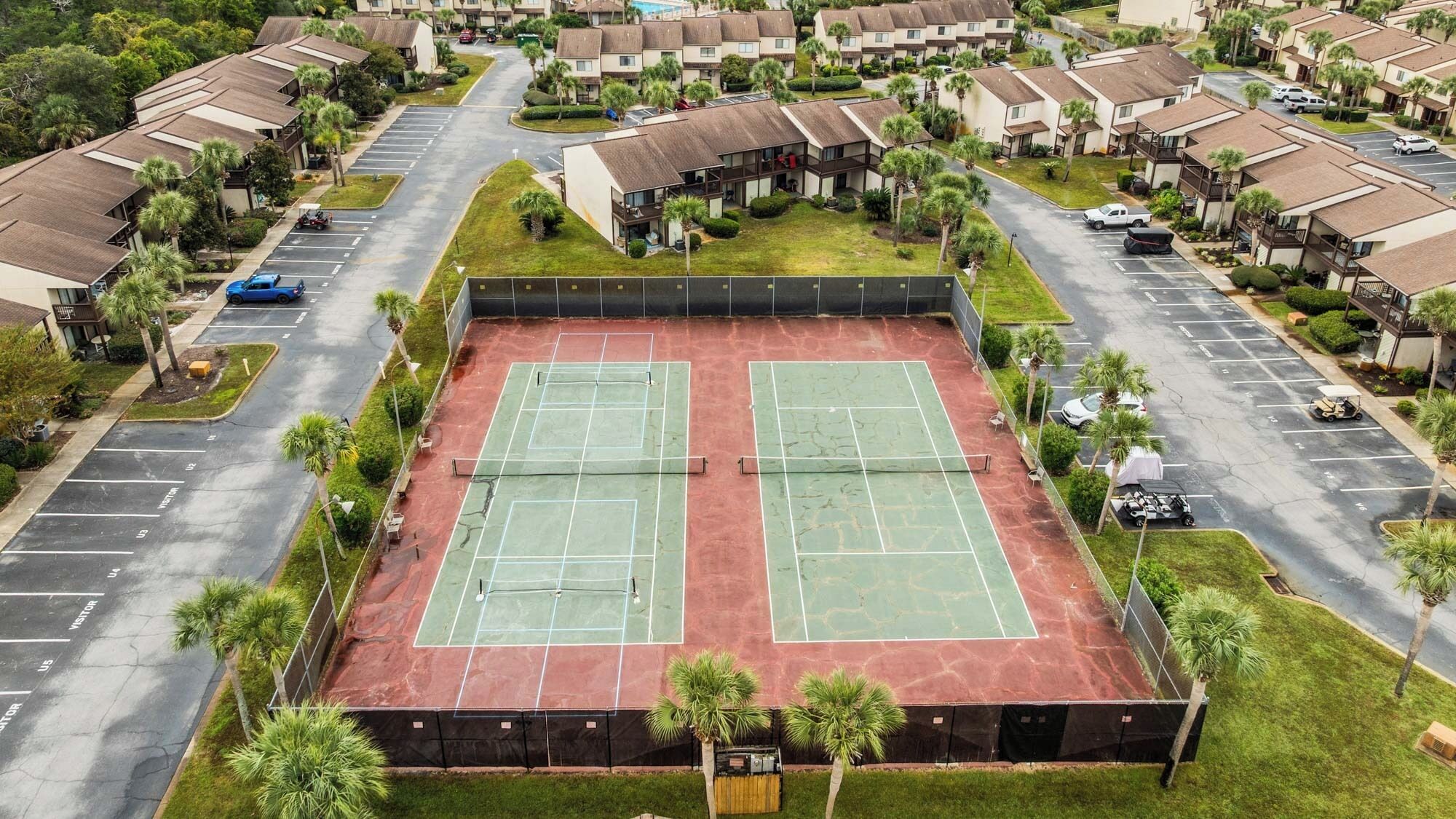 Un court de tennis entouré d’un parking, avec une rangée de maisons de ville en arrière-plan.