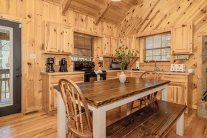 A wooden cabin interior with a kitchen featuring wooden cabinets, a dining table with chairs, a stove, and a microwave. - WhispPineR: Wooded View, Hot Tub, Wifi, Secluded, Game Room, Theater (Sevierville)