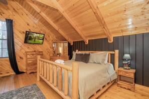 A wooden cabin bedroom with a bed, dresser, nightstand, and a television on the wall. - WhispPineR: Wooded View, Hot Tub, Wifi, Secluded, Game Room, Theater (Sevierville)