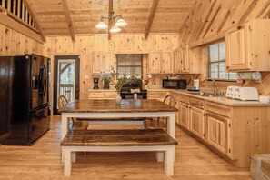 A wooden cabin interior with a dining table, benches, a refrigerator, a microwave, and wooden cabinets. - WhispPineR: Wooded View, Hot Tub, Wifi, Secluded, Game Room, Theater (Sevierville)