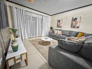 A living room with a grey sofa, a white coffee table, a grey rug, and a white vase with flowers. - Cozy Cedar Haven Fourways Mall City Escape (Fourways)