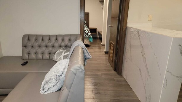 Un soggiorno moderno con un divano grigio, un telecomando sopra e un corridoio che porta al bagno.