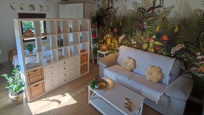 Una habitación con un sofá blanco, un ventilador de techo, una estantería, un mural tropical y una pequeña mesa con una planta.
