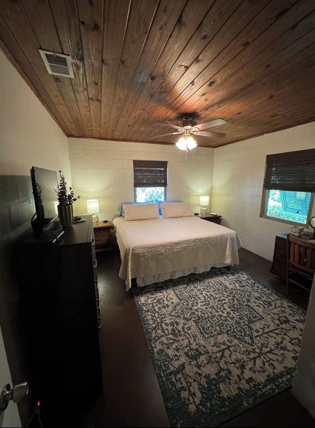 Une chambre avec un plafond en bois, un lit recouvert d’une courtepointe blanche, une table de chevet, un bureau, une chaise et une fenêtre avec des stores.