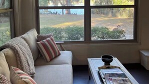 Una habitación con sofá, una mesa de centro con revistas y un ventanal con vistas a árboles y un muelle.