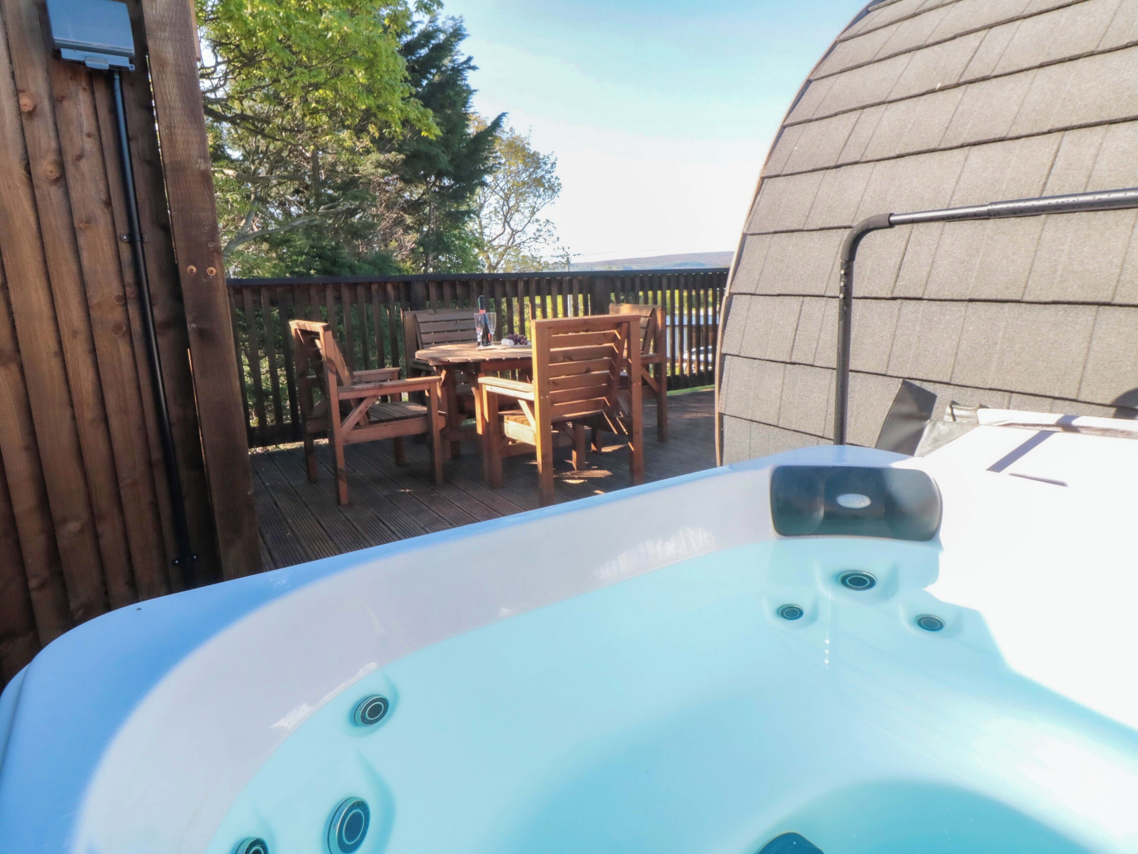 Ein Whirlpool auf einer Holzterrasse mit Tisch und Stühlen, umgeben von einem Holzzaun und Bäumen.