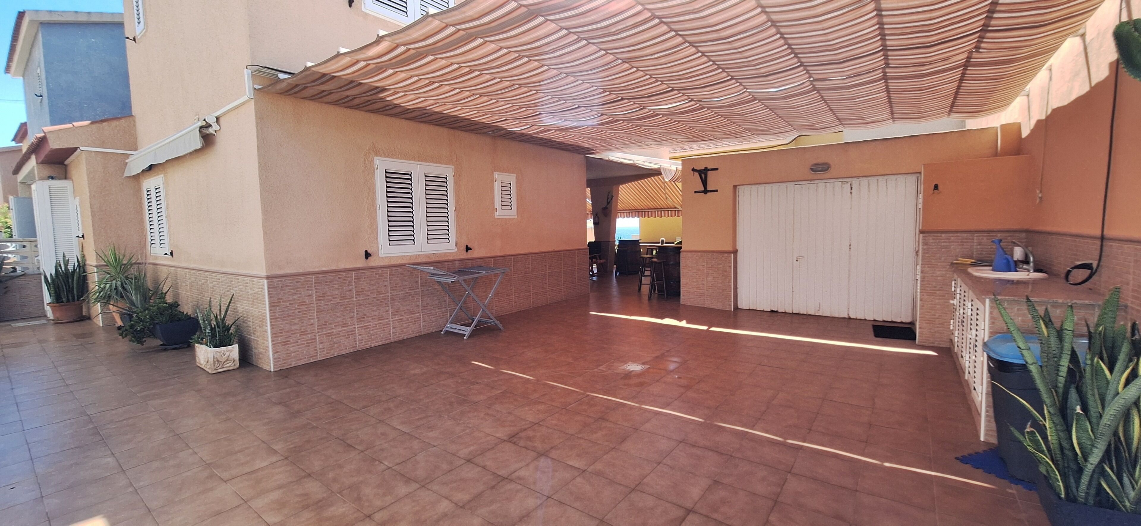 Área de varanda coberta com piso de azulejos, uma porta branca e plantas em vasos.