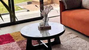 Un salon moderne avec une table basse en verre, un canapé, une plante et une vue sur un jardin grâce à de grandes fenêtres.