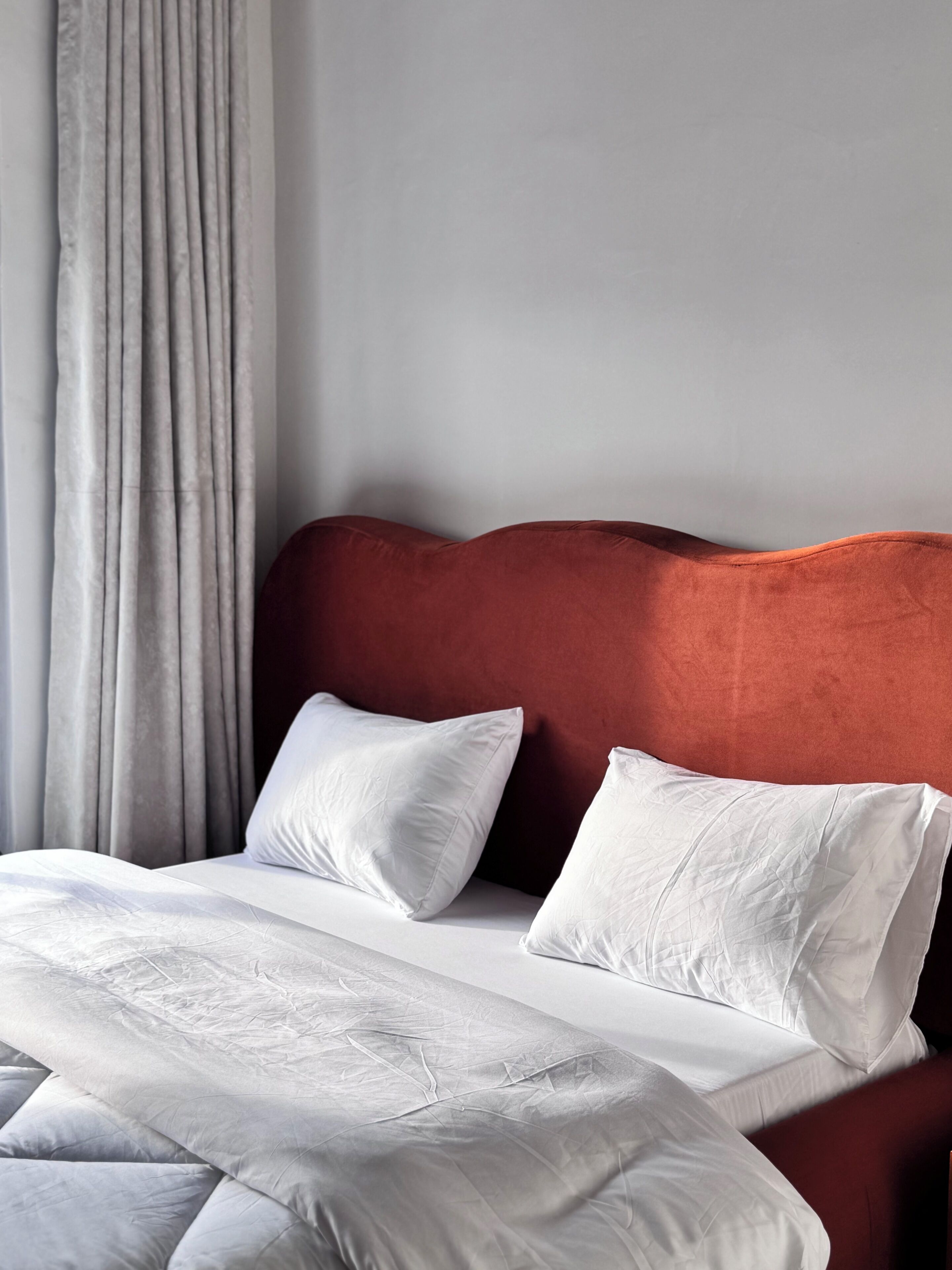 A bed with white bedding and pillows, a red headboard, and a grey curtain.
