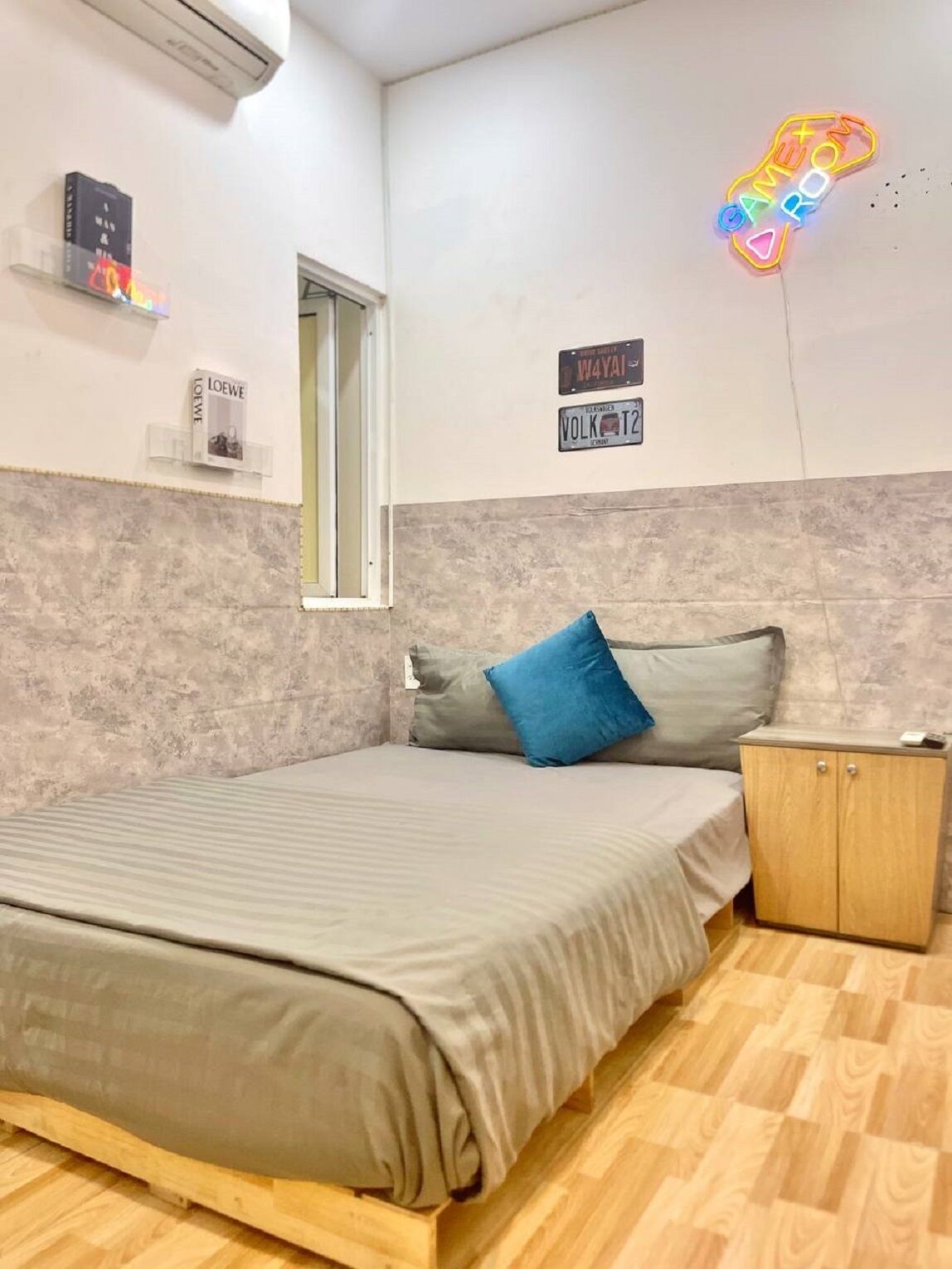A bedroom with a bed, a nightstand, a window, and wall decorations including license plates and a neon sign.