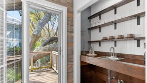 A kitchen with wooden shelves, a sink, and a view of a tree through a sliding glass door. - Live Oak Beach House (Isle of Palms)