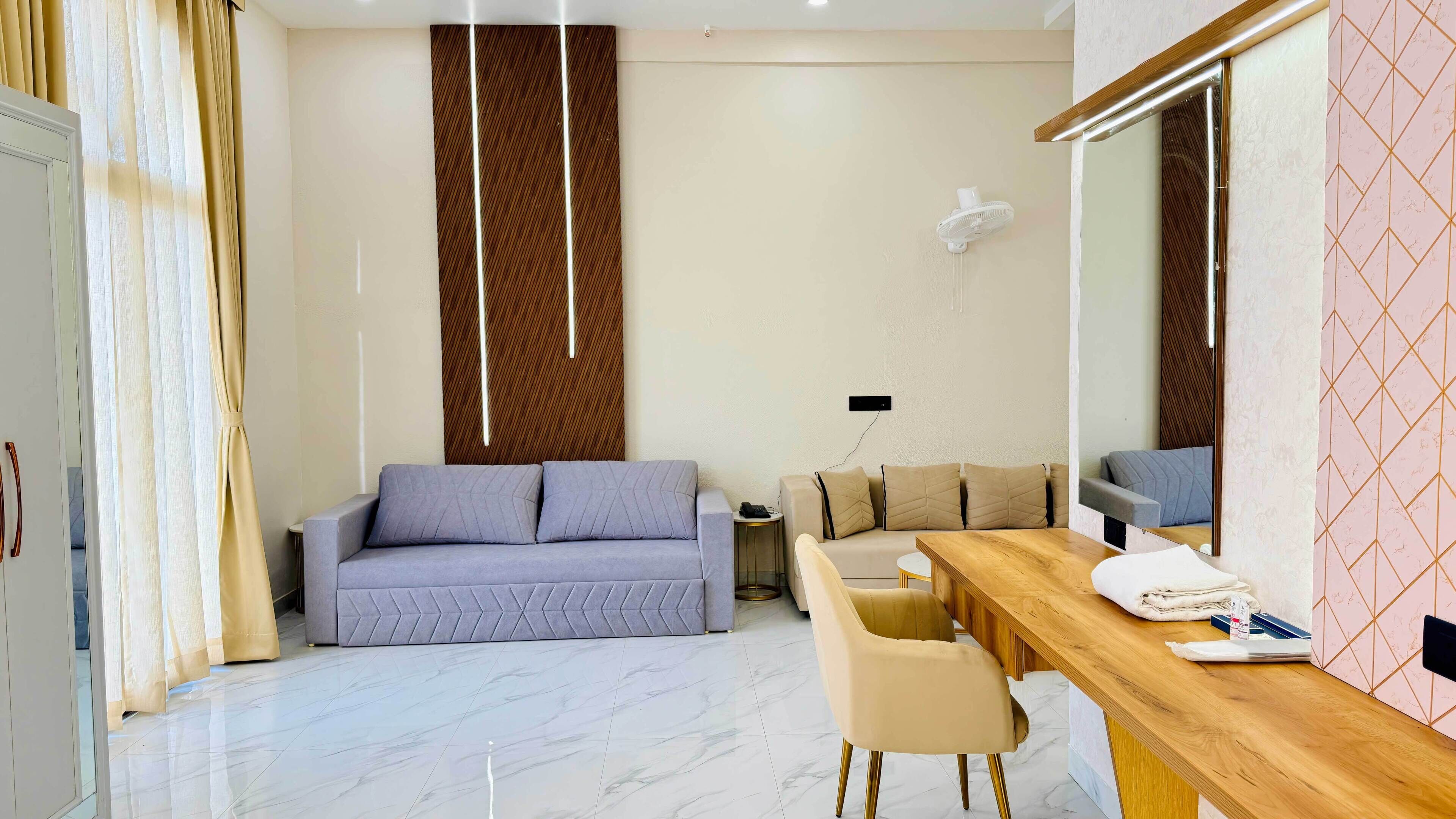 A modern hotel room with a sofa, a dining table, a chair, a mirror, and a fan.