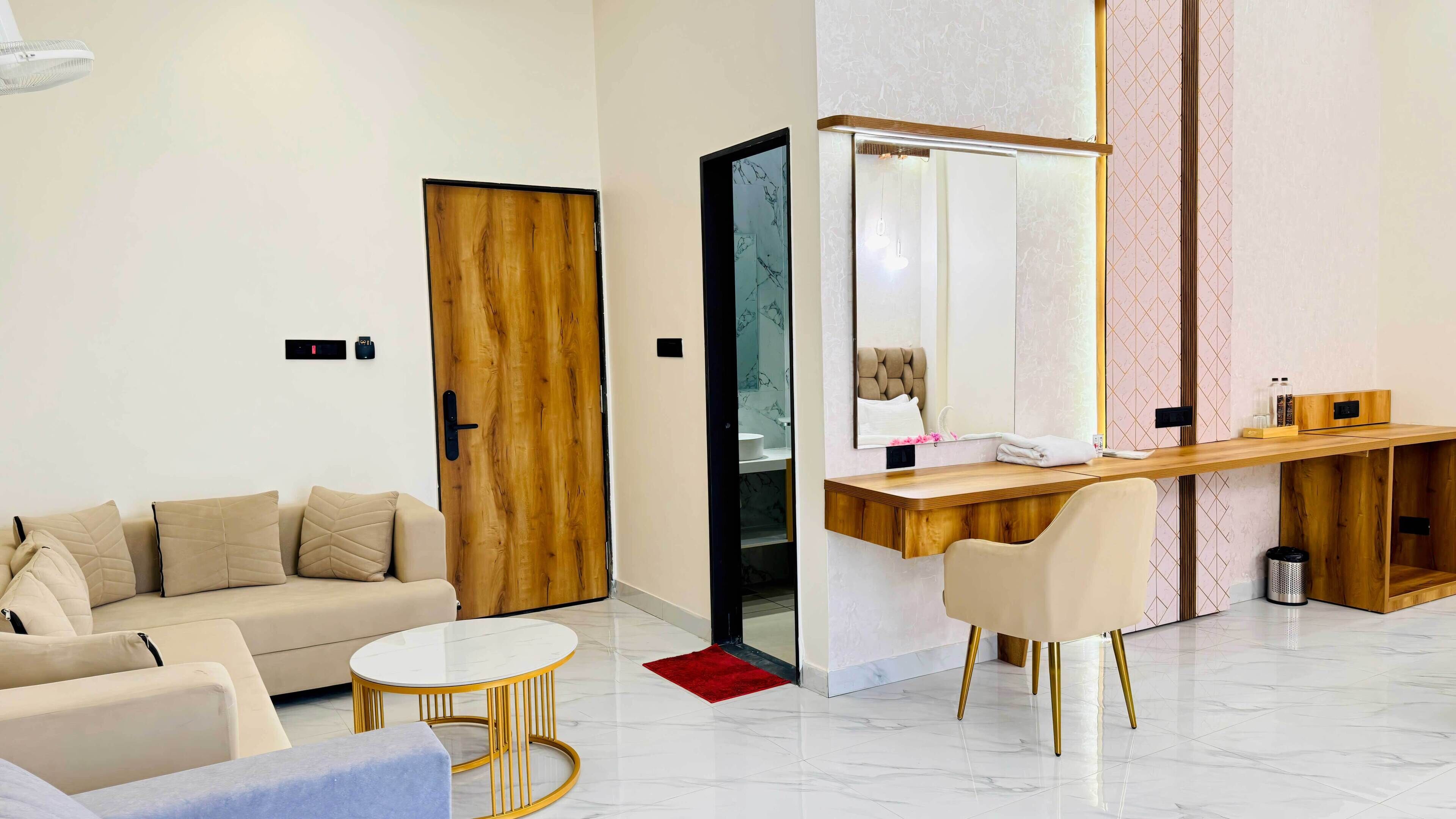 Une chambre d’hôtel moderne avec une porte en bois, un canapé, un bureau avec une chaise, une table ronde et un grand miroir.