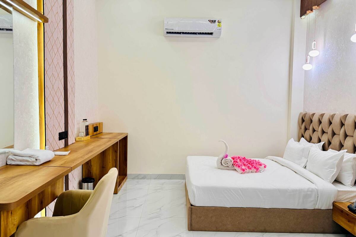 Habitación de hotel con escritorio de madera, cama con cabecera capitonada, espejo, unidad de aire acondicionado y mesita de noche con lámpara.