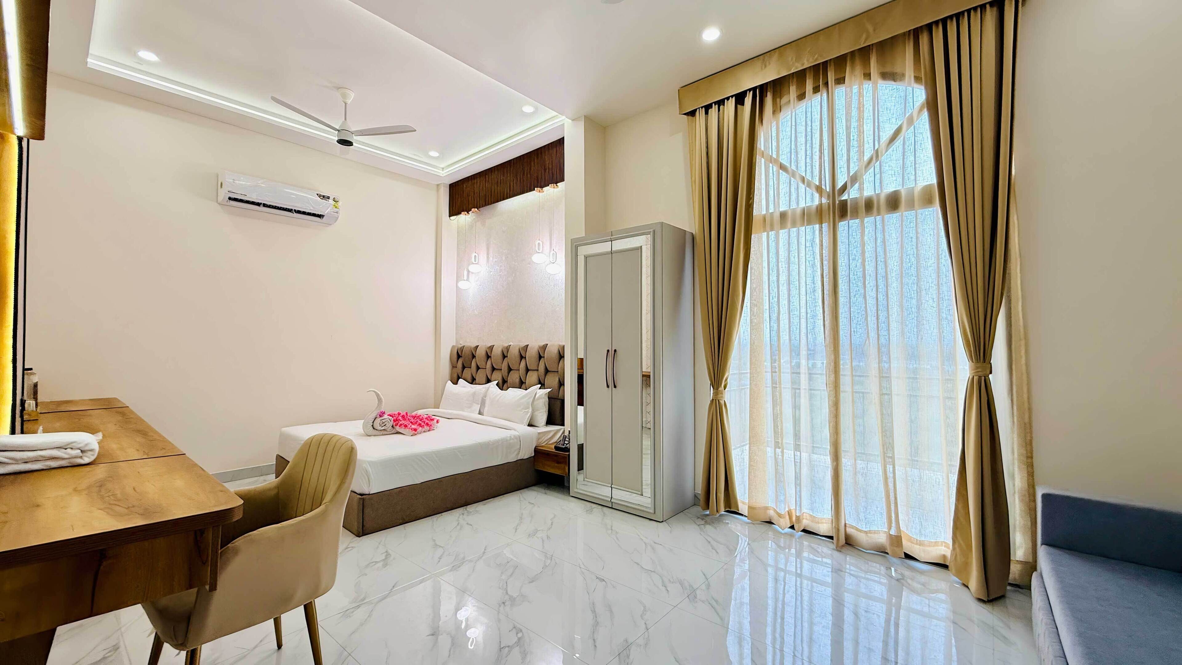 A hotel room with a bed, a wardrobe, a desk with a chair, and a large window with curtains.
