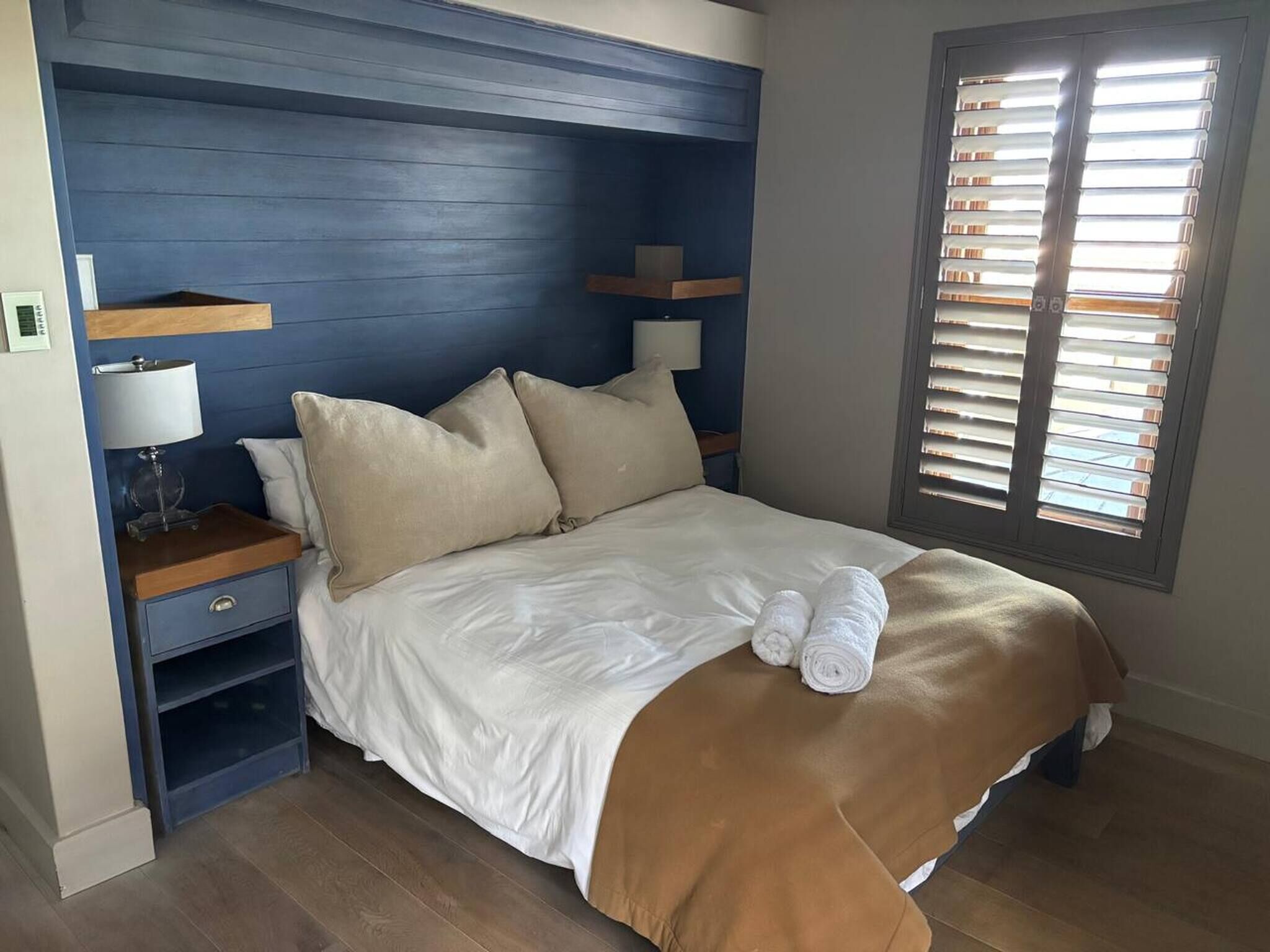 A bedroom with a bed, pillows, a nightstand, a lamp, and a window with blinds.