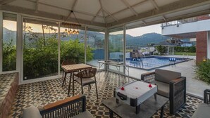 Une terrasse extérieure couverte avec une piscine, des sièges et une table, ainsi qu’un baby-foot.