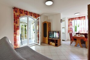 Ein Wohnzimmer mit einer Couch, einem Fernseher auf einem Holzboden, einem Esstisch mit Stühlen und einer Glastür zum Außenbereich.