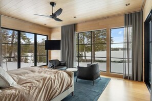 A modern bedroom with a large bed, a flat-screen TV, a ceiling fan, and large windows offering a view of trees.