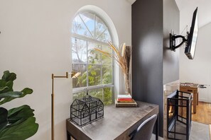 Ein Zimmer mit einem großen Bogenfenster, einer schwarzen Metall-TV-Wandhalterung, einem kleinen Tisch mit einem Buch, einer dekorativen Vase und einem hölzernen Tischfußballspiel.
