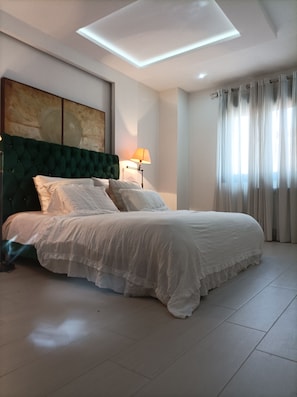 Une chambre à coucher avec un lit, une tête de lit, une lampe, un tableau et une fenêtre avec des rideaux.