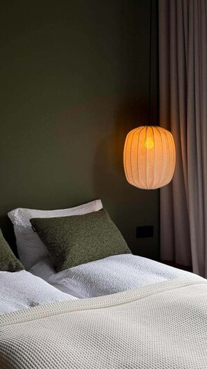 Un lit recouvert d’une couverture blanche à motifs, un oreiller vert et une lampe suspendue.