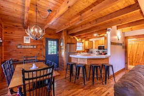 A wooden cabin interior with a dining area, kitchen, and bar stools. - Highwood- Mountain Views, Hot Tub, Arcade, Fire Pit, Foosball, Sleeps 12 (Boone)