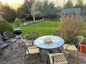 A patio with a round glass table and chairs, a barbecue grill, and a view of a well-maintained lawn and trees.