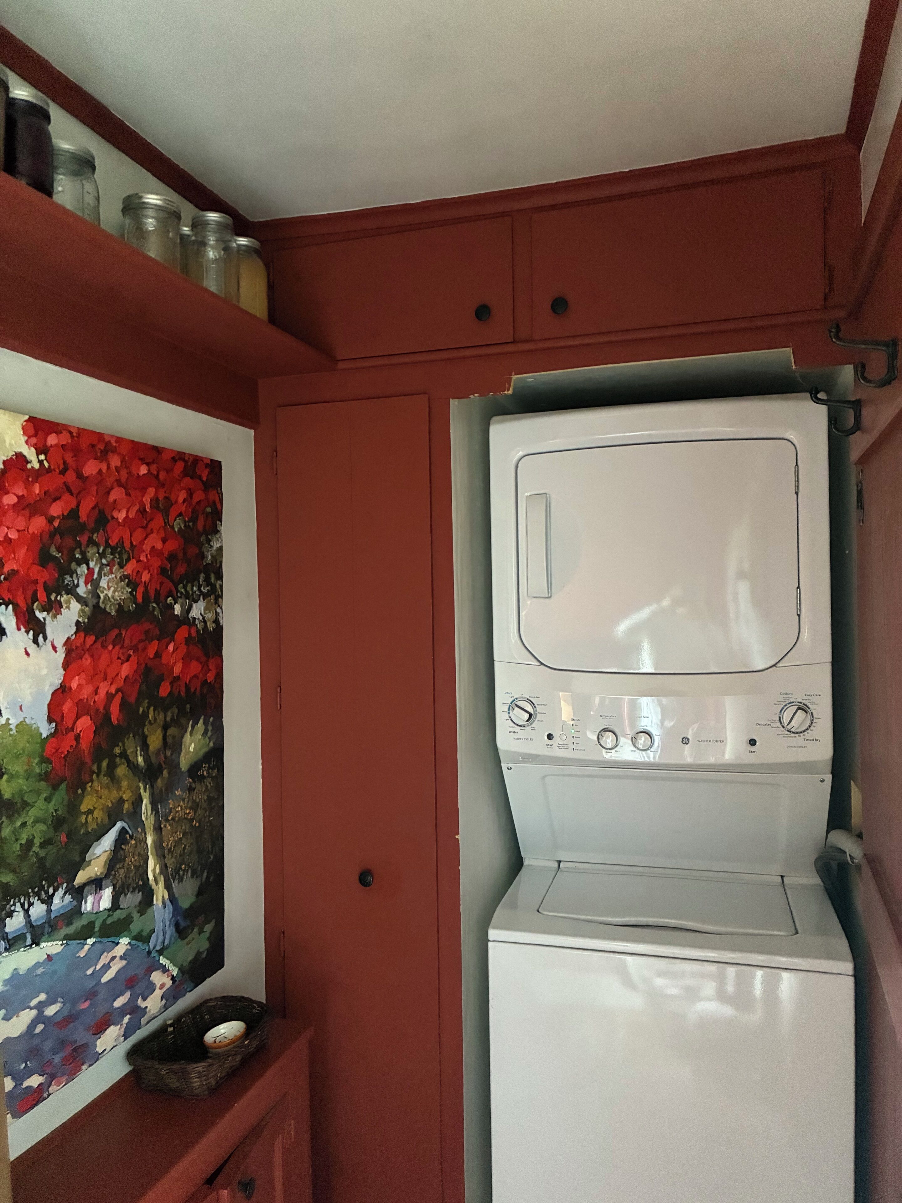 Une petite buanderie comprenant une machine à laver et un sèche-linge superposés, une étagère avec des bocaux et un tableau représentant un paysage.